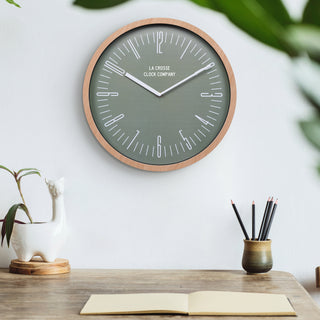 Lifestyle image of the 404-3041D 16-inch Ellwood Wall Clock, with Silent Sweep, shown hanging on a wall in a home office with a wood desk, desk accessories, and plants. The clock features a flush-mount light wood frame, a sage green dial, modern white numbers, and white metal hour and minute hands.