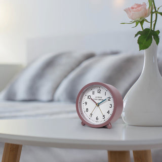 Lifestyle image of the 617-3816A Analog Quartz Alarm Clock sitting on a white bedside table with a white vase filled with pink roses with a bed in the background. The alarm clock features a maroon case and a white dial with black numbers, a maroon hour hand, a blue minute hand, and brass second hand and alarm indicator.