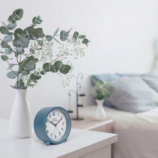 Lifestyle image of the 617-3816B Analog Quartz Alarm Clock sitting on a white bedside table with a white vase filled with foilage with a bed in the background. The alarm clock features a blue case and a white dial with black numbers, a blue hour hand, a maroon minute hand, and brass second hand and alarm indicator.