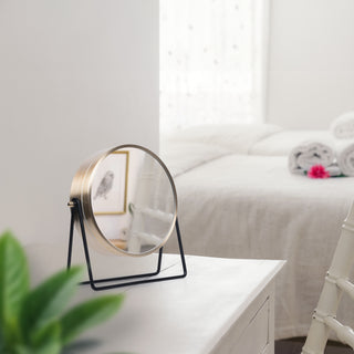 404-3719 A round vanity mirror with a gold-toned metal frame and black stand, displayed on a white bedside table in a softly lit bedroom.