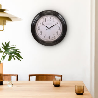 Lifestyle image of the 404-2658 23-inch Maxwell Wall Clock - Arabic Numerals, the clock is shown hanging on a white wall in a dining room with a mid-century modern table and chairs. The clock features a parchment paper-inspired dial with distressed numbers, and decorative metal hands in a dark antiqued bronze frame.