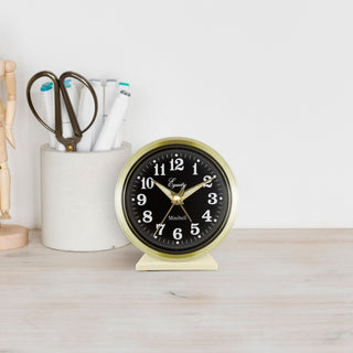 Lifestyle image of the 12020 Analog Keywind Alarm Clock featuring a brass case, black dial with white numbers, and brass hands. The clock is shown sitting on a light wood desk with desk accessories.