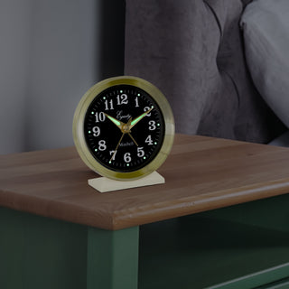 Lifestyle image of the 12020 Analog Keywind Alarm Clock featuring a brass case, black dial with white numbers, and brass hands sitting on a nightstand in a dark room.