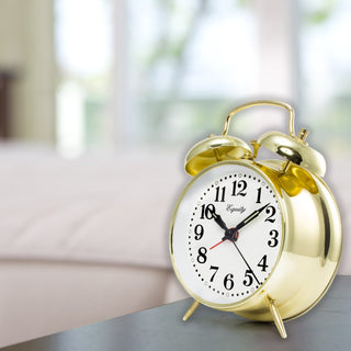 A shiny gold twin-bell alarm clock with a white face and black numbers, sitting on a dark tabletop with a softly blurred indoor background.