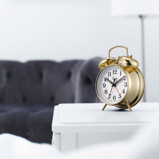 Lifestyle image of the 13012 Metal Key Wind Alarm Clock - Gold, shown on a white side table with a dark gray tufted fabric couch in the background. The classic twin-bell alarm clock features a polished gold finish metal body, white clock face, black numerals and hands, and a red second hand. The brand name 'Equity' is printed on the face.