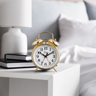 Lifestyle image of the 13012 Metal Key Wind Alarm Clock - Gold shown on a white nightstand with a bed in the background. The classic twin-bell alarm clock features a polished gold finish metal body, white clock face, black numerals and hands, and a red second hand. The brand name 'Equity' is printed on the face.