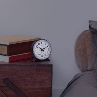 A round black analog alarm clock with a white face and black numbers sits on a dark wooden bedside table next to stacked books and a bed.