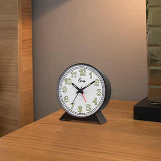 A round analog desk clock with a black metal frame and base, a white face, green numerals, and black hands with a red second hand, shown on a wooden tabletop in an indoor setting.