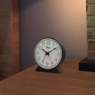A round analog desk clock with a black metal base and a white face, featuring green numerals and black hands with a red second hand. The clock is shown on a wooden tabletop in a warm, dimly lit room.