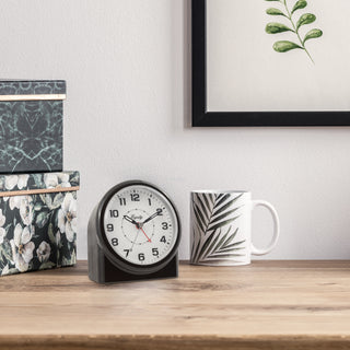 Black clock on a wooden surface with decorative items including a mug and floral boxes.