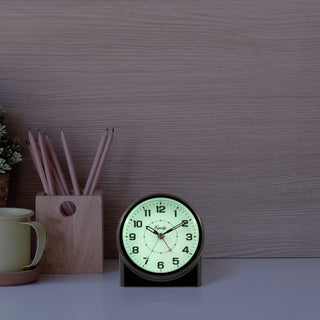 A round analog desk clock with a black frame and a glowing pale green face, shown on a tabletop against a wood-grain wall. A wooden pencil holder and part of a mug are visible to the left.