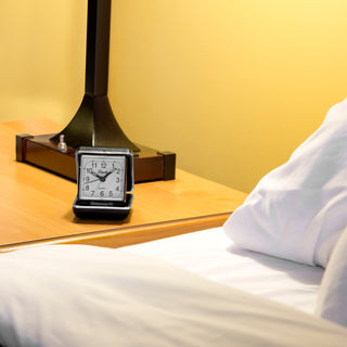 A small square analog alarm clock with a black frame and white face sits on a wooden bedside table next to a bed with white bedding and a lamp base in the background.