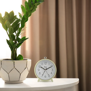 A round vintage-style alarm clock with a pale green frame and white face sits on a white table next to a potted green plant, with beige curtains in the background.