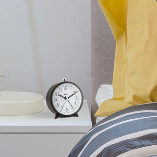 A round analog alarm clock with a dark gray frame and white face sits on a white bedside table next to a bed with yellow curtains and blue-and-white striped bedding.