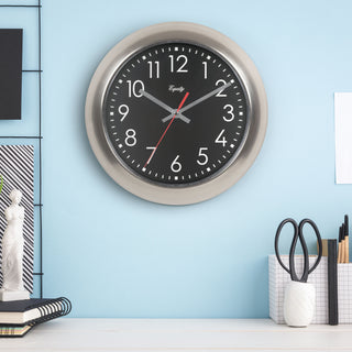 Lifestyle image of the 20802 11-inch Owen Metal Wall Clock with Silent Sweep shown hanging on a blue wall above a white desk with desk accessories, notebooks, and a white Venus de Milo miniature statue. The clock features a black dial with white numbers, silver hour and minute hands, a red second hand, and a brushed titanium finished metal frame.