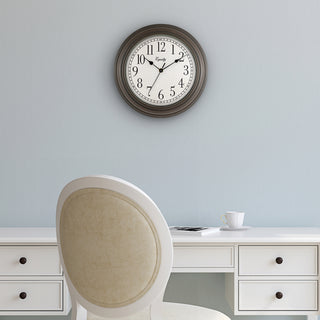 Lifestyle image of the 20898 Cora 12-inch Wall Clock hanging on a light blue wall above a white desk and chair. The clock features a classic dial, black numbers, seconds indicator ring, and an oil-rubbed bronze frame