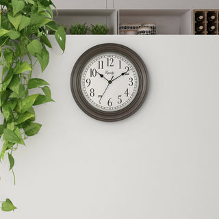 Lifestlye image of the 20898 Cora 12-inch Wall Clock hanging on a white wall with shelves above the clock and a green plant. The clock features a classic dial, black numbers, seconds indicator ring, and an oil-rubbed bronze frame