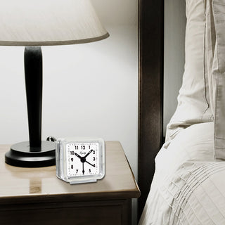 A small square analog alarm clock with a clear outer frame and white face sits on a wooden bedside table next to a black table lamp and a bed with white linens.