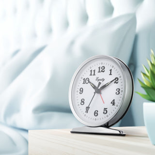 Lifestyle image of the 24014 Analog Wind-Up Loud Bell Alarm Clock shown on white nightstand next to a bed with blue bedding in the background. The alarm clock features a chrome case with a white dial, black numbers and hands, luminous hour and minute hands, luminous hour dots, and a red alarm indicator. The 'Equity' brand logo is printed in black script on the clock face.