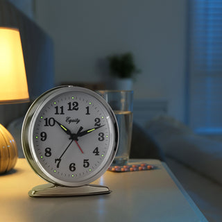 Lifestyle image of the 24014 Analog Wind-Up Loud Bell Alarm Clock shown on a nightstand in a dark bedroom with a lamp and a glass of water in the background. The alarm clock features a chrome case with a white dial, black numbers and hands, luminous hour and minute hands, luminous hour dots, and a red alarm indicator. The 'Equity' brand logo is printed in black script on the clock face.