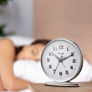 Lifestyle image of the 24014 Analog Wind-Up Loud Bell Alarm Clock shown on a wood nightstand in the foreground with a women sleeping in bed in the backgrond. The alarm clock features a chrome case with a white dial, black numbers and hands, luminous hour and minute hands, luminous hour dots, and a red alarm indicator. The 'Equity' brand logo is printed in black script on the clock face.
