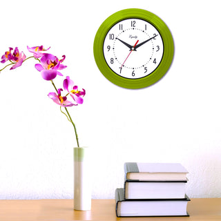 Lifestyle image of the 25016 8-inch Basic Wall Clock by Equity - Lime Green shown hanging on a white wall above a wood table with a vase of hot pink orchids. The clock features a fluted lime green frame, a white dial, black numbers, black hour and minute hands, and a red second hand.