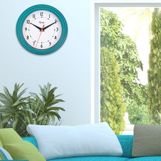 Lifestyle image of the 25020 8-inch Basic Wall Clock by Equity - Teal Blue, hanging on a light blue wall next to an entryway, above a teal couch, and plants. The clock features a fluted teal blue frame, a white dial, black numbers, black hour and minute hands, and a red second hand.
