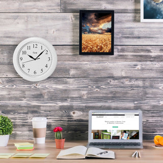 Lifestyle image of the 25201 10-inch Basics Wall Clock - White shown hanging on a weathered wood paneled wall above a desk with a laptop, notebook, post-its, a cactus, and a coffee cup. The clock features a white frame with a white dial, black numbers, and black metal hands.