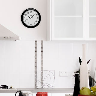 Lifestyle image of the 25203 10-inch Basics Wall Clock - Black shown hanging on a white tiled kitchen wall above a gray countertop. The clock features a black frame with a white dial, black numbers, and black metal hands.