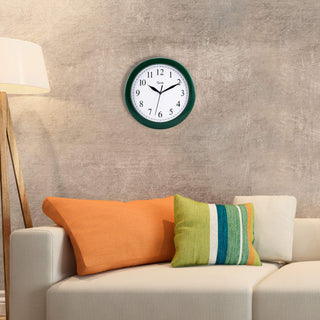 Lifestyle image of the 25205 10-inch Basics Wall Clock - Hunter Green shown hanging on a tan wall above a cream couch with orange and multi-colored striped accent pillows. The clock features a hunter green frame with a white dial, black numbers, and black metal hands.