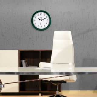 Lifestyle image of the 25205 10-inch Basics Wall Clock - Hunter Green shown hanging on a gray wall about a bookshelf in an office setting. The clock features a hunter green frame with a white dial, black numbers, and black metal hands.