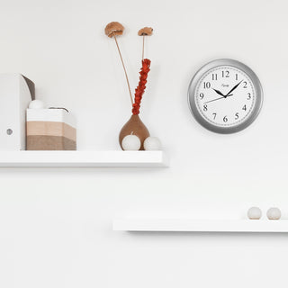 Lifestyle image of the 25206 10-inch Basics Wall Clock - Silver shown hanging on a white wall above white shelves with candles and a vase. The clock features a silver frame with a white dial, black numbers, and black metal hands.