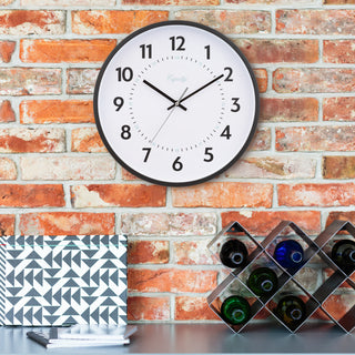 Lifestyle image of the 25509 14-inch Basics Commercial Wall Clock hanging on a sienna brick wall above a gray countertop with notepads and a wine rack. The clock features 24-hour markings in light blue and black numbers on a white dial with black hour and minute hands, and a silver second hand surrounded by a black flush-mount frame.