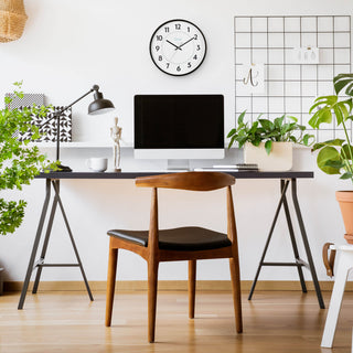Lifestyle image of the 25509 14-inch Basics Commercial Wall Clock hanging on a white wall in a home office environment with a desk, computer, plants, and a chair. The clock features 24-hour markings in light blue and black numbers on a white dial with black hour and minute hands, and a silver second hand surrounded by a black flush-mount frame.