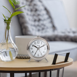 A silver analog alarm clock with a white face and black numerals sits on a light wood side table in a cozy living room setting, next to a glass vase with green leaves and a white mug.