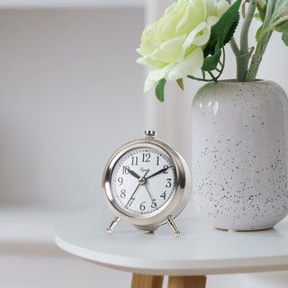 A silver analog alarm clock with a white face and black numerals sits on a white table next to a speckled gray ceramic vase holding pale green flowers.