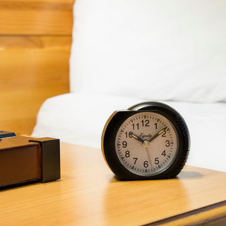 A round analog alarm clock with a black body and white face sits on a wooden bedside table next to a leather suitcase, with a blurred bed in the background.