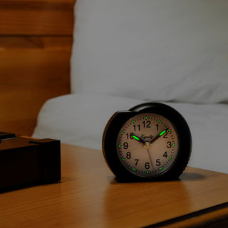 A round black alarm clock with glowing green numerals and hands sits on a wooden bedside table in front of a bed with white bedding.