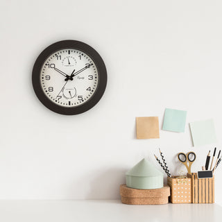 Lifestyle image of the 29005 10-inch Waylon indoor outdoor wall clock with temperature and humidity, featuring a linen design dial with a dark brown frame, stylized cut-out hour and minute hands, and a second hand hanging on a white wall above a desk with office supplies in soft neutral tones