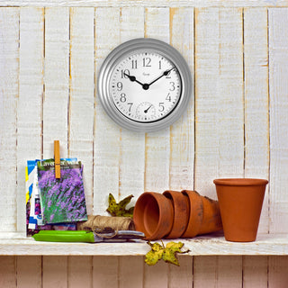 A round wall clock with a silver frame and white face hangs on a white wood-paneled wall above a shelf with gardening props, including terracotta pots, shears, twine, and a lavender photo.