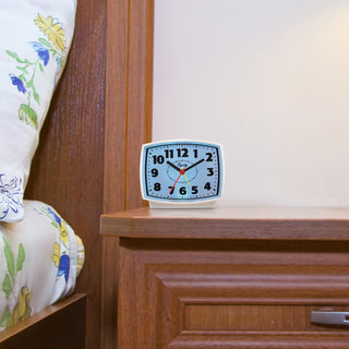 Lifestyle image of the 33100 Electric Analog Alarm Clock with Backlight sitting on a nightstand next to a bed with yellow and blue floral bedding, showing the blue backlight illuminated.