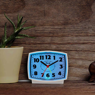 Lifestyle image of the 33100 Electric Analog Alarm Clock with Backlight sitting on a dark stained bookshelf next to a cactus, showing the blue backlight illuminated in a darkened room.