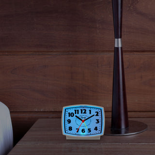 Lifestyle image of the 33100 Electric Analog Alarm Clock with Backlight sitting on a nightstand next to a lamp with dark paneled walls. The alarm clock has the blue backlight illuminated in a darkened room.
