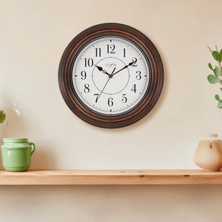 Lifestyle image of the 404-2630W Evelyn wall clock in bronze with a white dial and black stylized Arabic numbers hanging on a beige wall above a pine shelf with a green ceramic creamer and tan teapot with a hint of plants