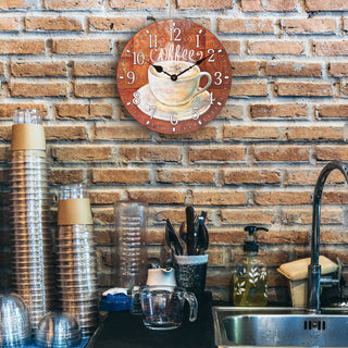 Lifestyle image of the 404-2631C 12-inch Coffee Wall Clock hanging on a brick wall above a coffee station. The clock features an image of an off-white steaming coffee cup and white numbers on a rusty brown background with no lens and black metal hands.