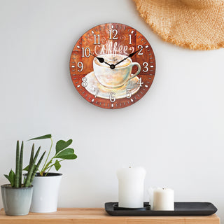 Lifestyle image of the 404-2631C 12-inch Coffee Wall Clock shown hanging on a white wall above a wood shelf with plants and candles. The clock features an image of an off-white steaming coffee cup and white numbers on a rusty brown background with no lens and black metal hands.