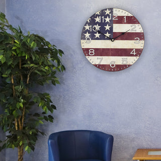 Lifestyle image of the 404-2631F 12-inch Flag Wall Clock hanging on a blue textured wall. The clock features an image of the American flag with white numbers, with no lens, and black metal hands.