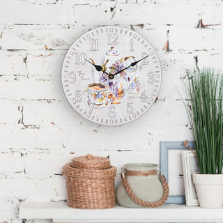 Lifestyle image of the 404-2631T 12-inch Teatime Wall Clock shown hangin on white-washed brick wall above a mantele with a woven container, a gree container, and a plant. The clock features an image of a floral teapot and teacup on a light-textured background, with the text "Tea Time," white numbers, black metal hands, and an open face design.