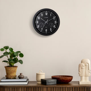 Lifestyle image of the 404-2631 12-inch Inkwell Wall Clock featuring a black flush mount frame surrounding a black dial with white numbers, thermometer that reads both Fahrenheit and Celsius, and a Humidity gauge, with silver metal hour, minute, and second hands hanging on a tan wall above a side table with a wood bowl, plant, ivory sculpture, and vase.