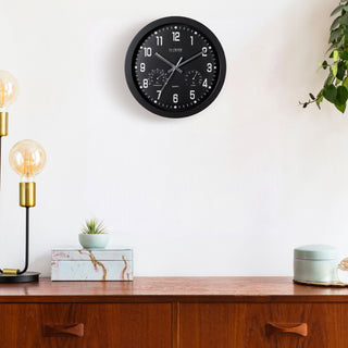 Lifestyle image of the 404-2631 12-inch Inkwell Wall Clock featuring a black flush mount frame surrounding a black dial with white numbers, thermometer that reads both Fahrenheit and Celsius, and a Humidity gauge, with silver metal hour, minute, and second hands hanging on a light colored wall above a buffet table